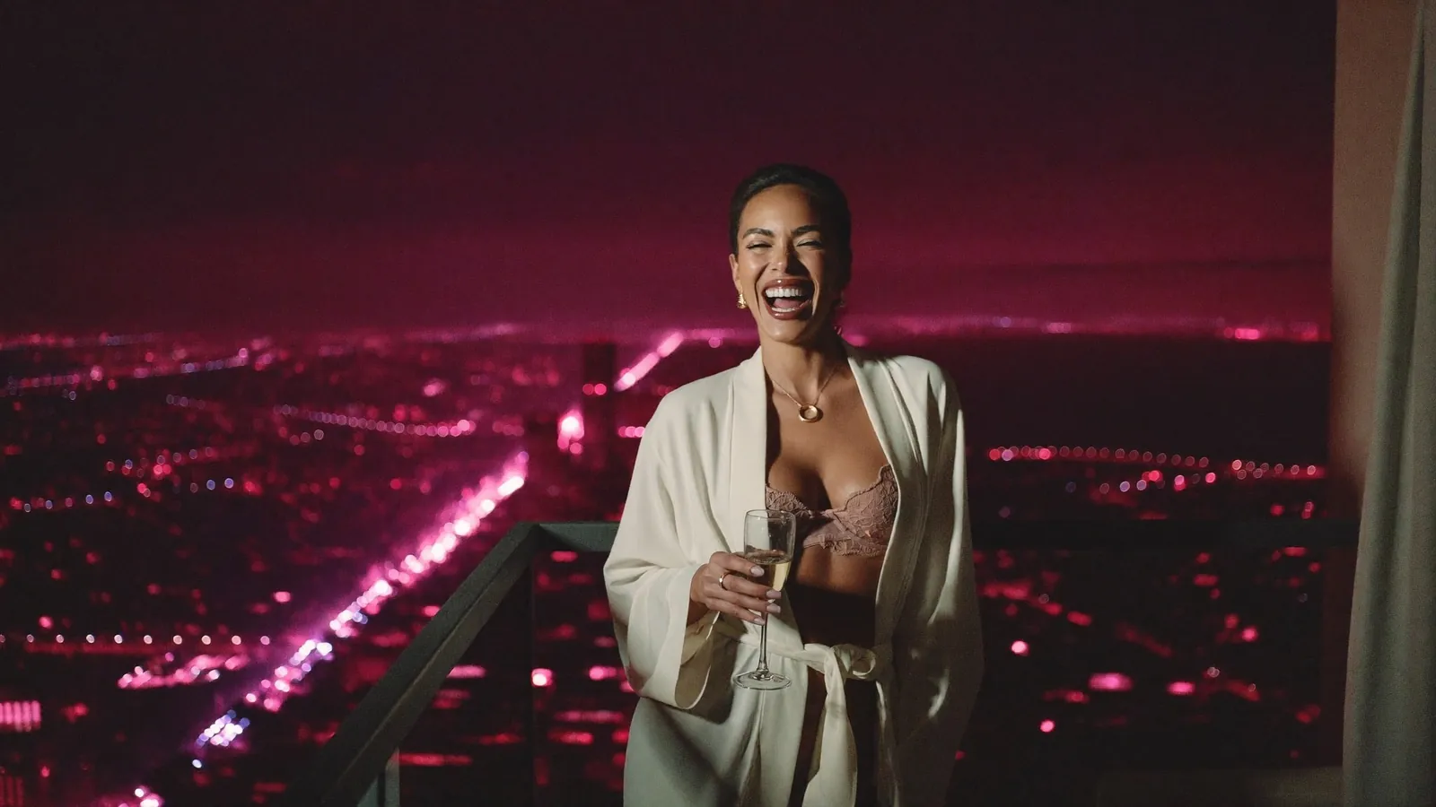 Cam model in a silk robe holding champagne on a city-view balcony