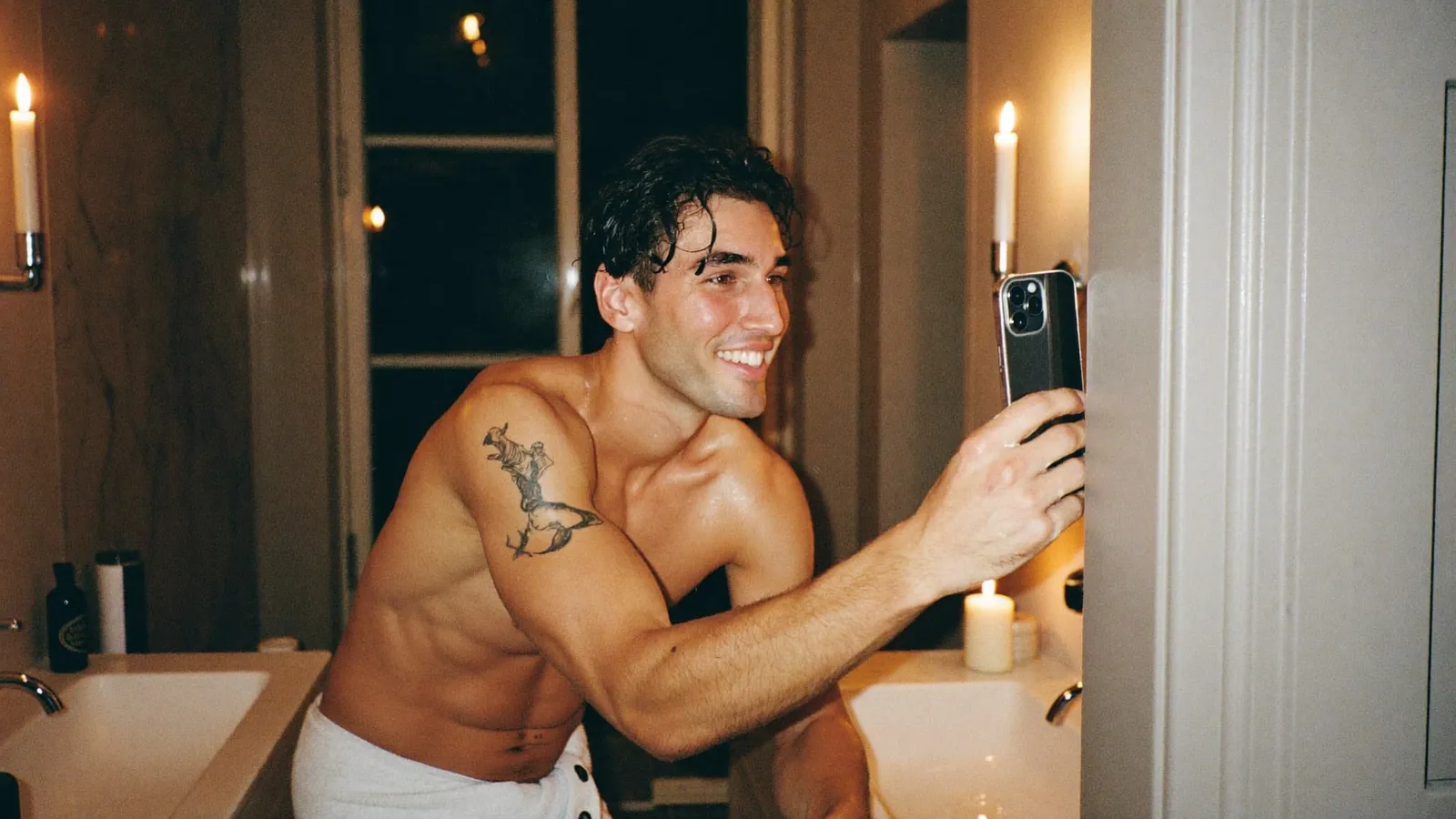 Tattooed cam model in a towel taking a selfie in a candlelit bathroom
