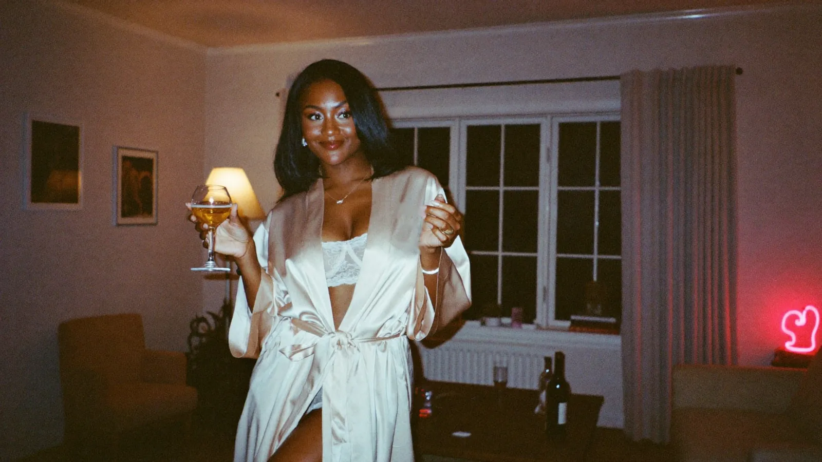 Smiling cam model in an ivory satin robe and white lace holding a glass of wine at home