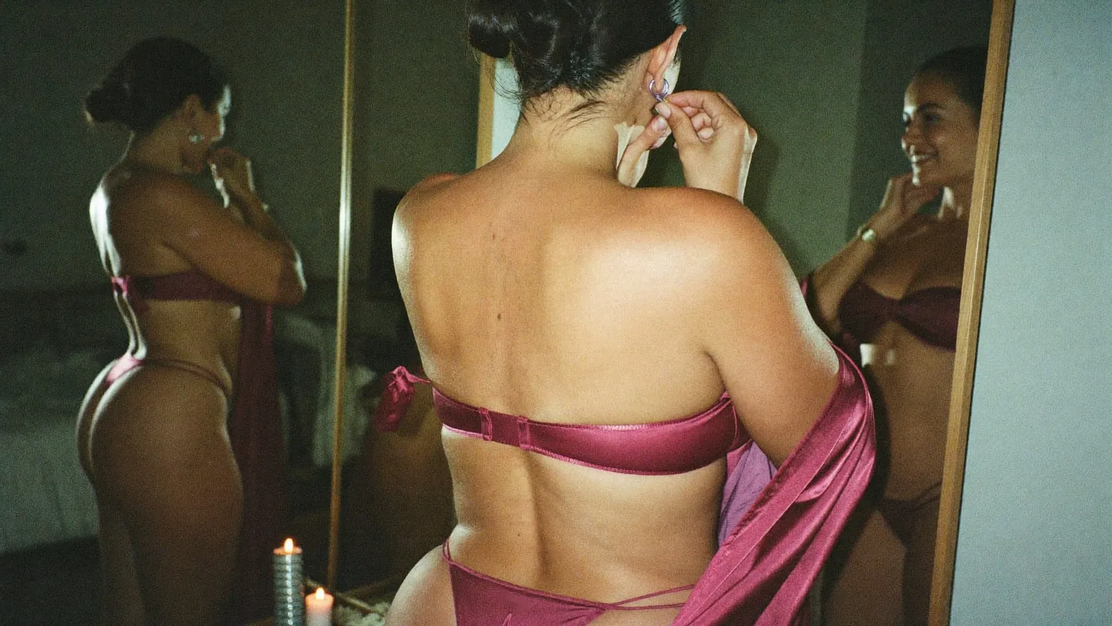 Cam model in plum satin lingerie putting on an earring between two mirrors