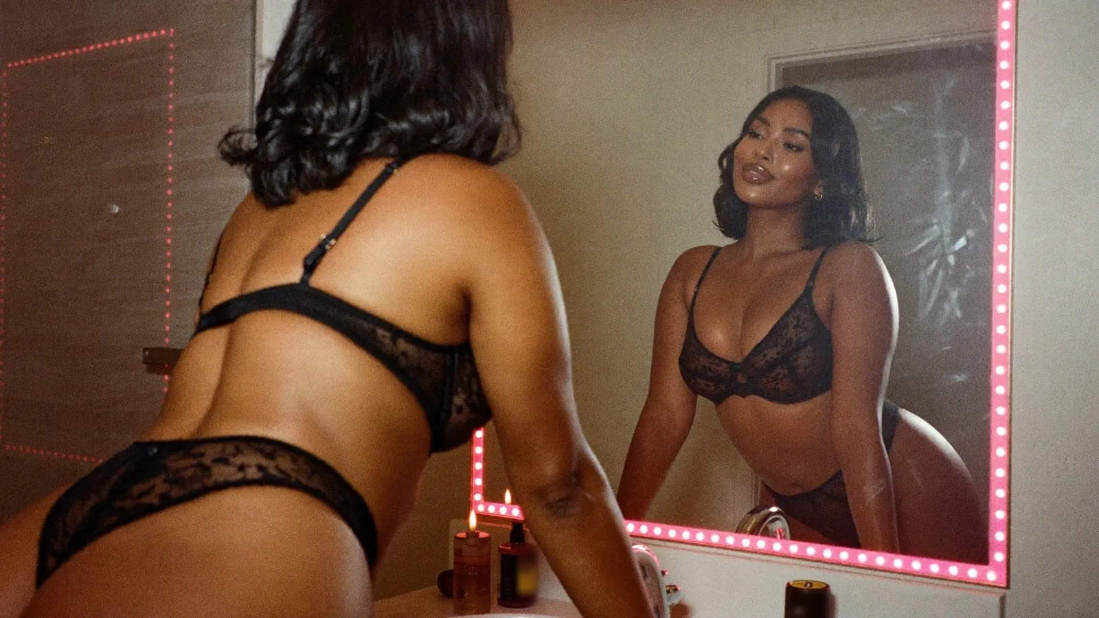 Cam model in black lace at a mirror lined with pink lights