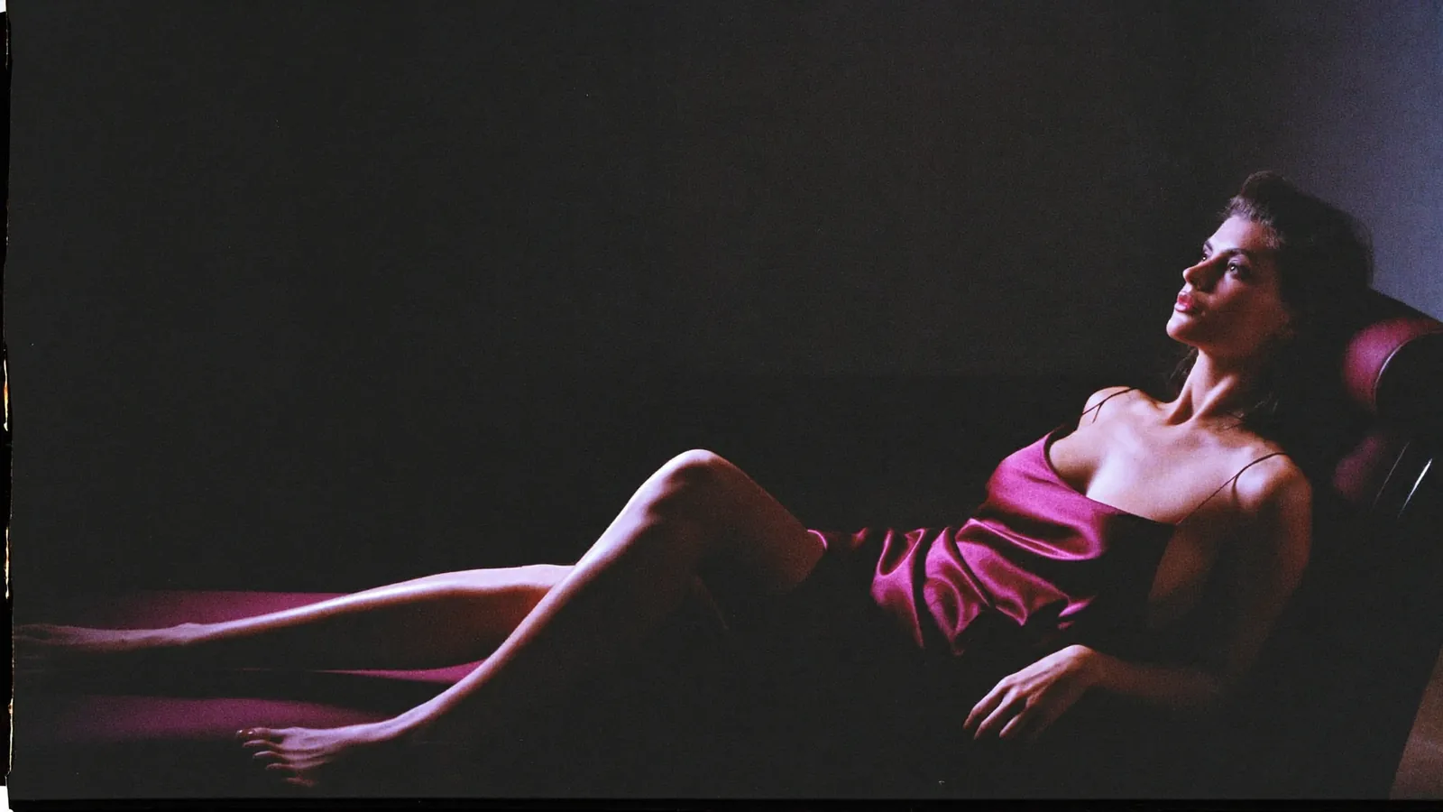 Trans model in a ruby satin slip reclining on a chaise in a dark room