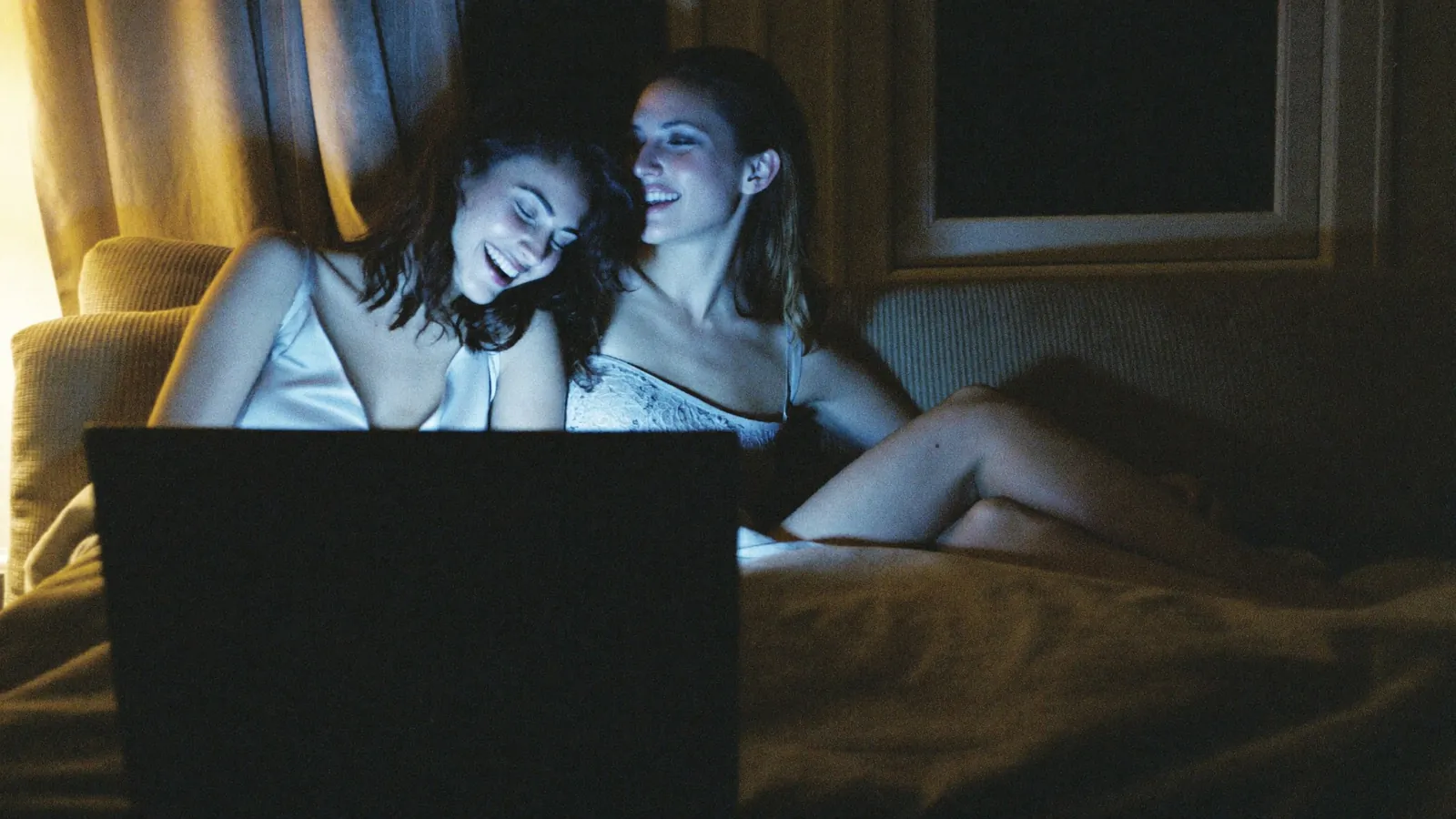 Two women in satin slips laughing together in front of a laptop at night