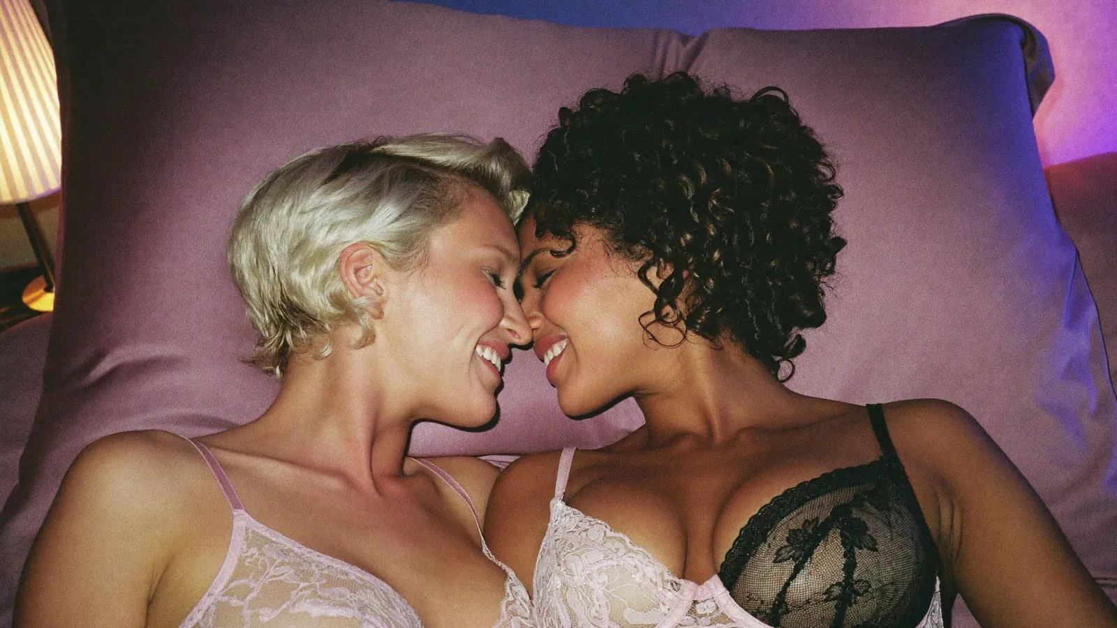 Two women in lace lingerie smiling at each other on a pillow