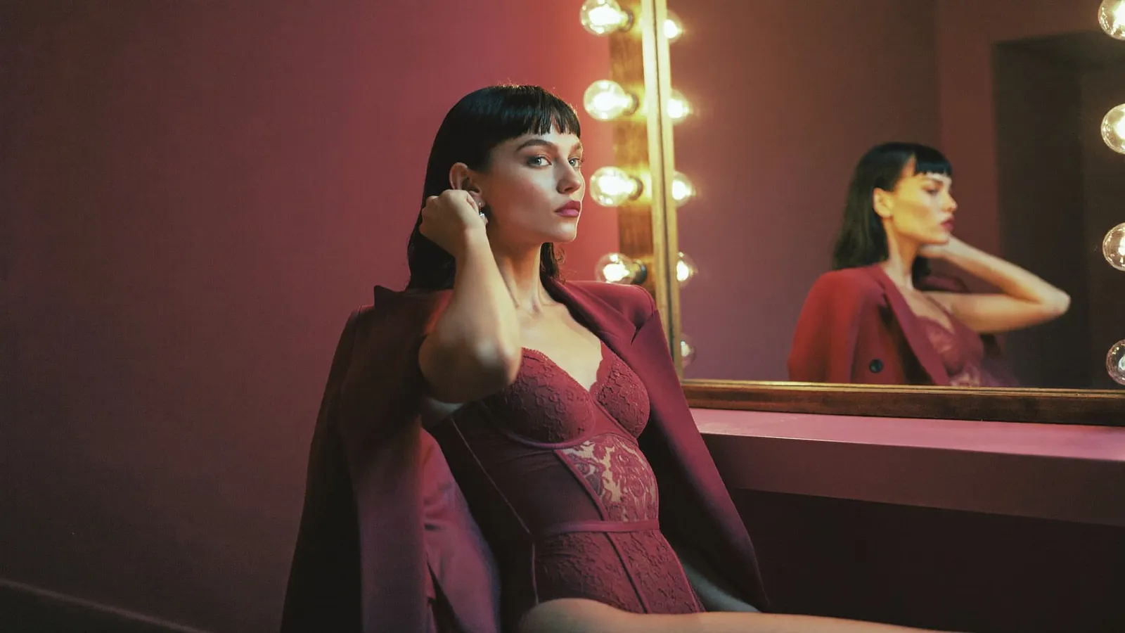 Cam model in burgundy lingerie at a lit vanity mirror